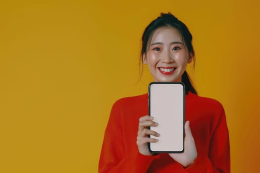 Asian woman in red sweater holding a blank screen smartphone with toothy smile on yellow background AIG51A.