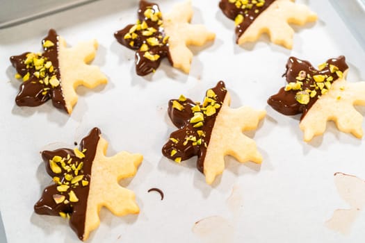 Crafting star-shaped holiday cookies dipped in chocolate and sprinkled with crushed pistachios, arranged on parchment paper.