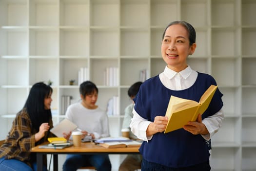 Positive senior professor holding book standing in class and looking away.