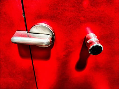 Close-up of a shiny metallic door handle and a latch set against a crimson backdrop. Close-Up of Chrome Handle and Latch on a Vibrant Red Door