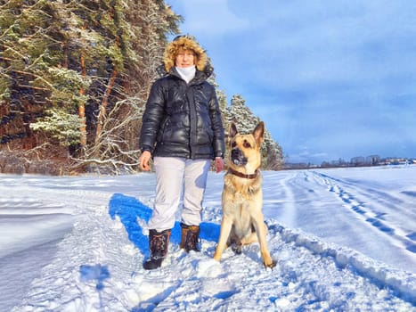 Adult girl or mature lady walking with shepherd dog, taking selfies in winter nature landscape. Middle aged woman and big shepherd dog on wooden bridge in cold day. Friendship, love, communication