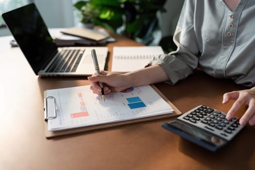 A woman is writing on a piece of paper with a pen. She is sitting at a desk with a laptop and a calculator. The scene suggests that she is working on some financial or business-related tasks