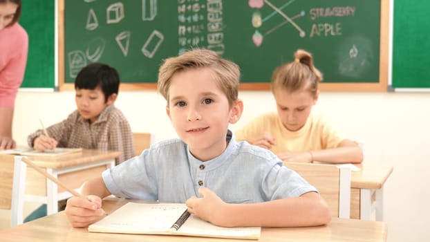 Attractive caucasian boy looking at camera while studying math at school. Diverse cute student doing test while teacher asking asian child question and point at paper in front of blackboard. Pedagogy.