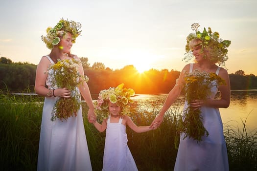 Ivan Kupala Celebration. Three Girls With Floral Wreaths by the River at Sunset. Family clad in white dresses celebrate Ivan Kupala by river at dusk.