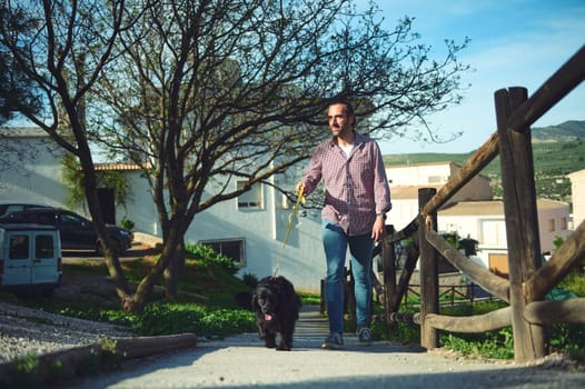 Smiling happy Caucasian man in checkered shirt and blue denim jeans, enjoys time with hid pet, walking his dog, a black cocker spaniel on leash on the nature. People and animals. Full length portrait