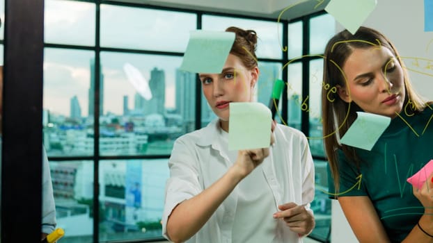 Professional business team brainstorm, sharing, thinking idea while skilled male leader write on sticky notes at glass wall. Skilled manager placed business plan on glass wall at office. Tracery