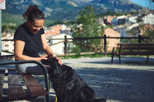 Woman sitting on a bench in the mountains, stroking her dog, smiling enjoying a happy time with her pet in the summer nature. Playing pets, people and animals concept. The dog is my best companion.