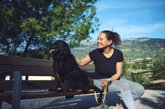 Cute black cocker spaniel dog sitting near his family on the bench in mountains nature. Beautiful woman stroking a her pat while walking it outdoors. People, domestic animals and nature
