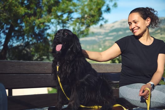 Beautiful woman sitting on city bench with her dog during walk in the morning. Smiling pretty woman stroking her young black cocker spaniel pet, sitting on a bench in the mountains nature