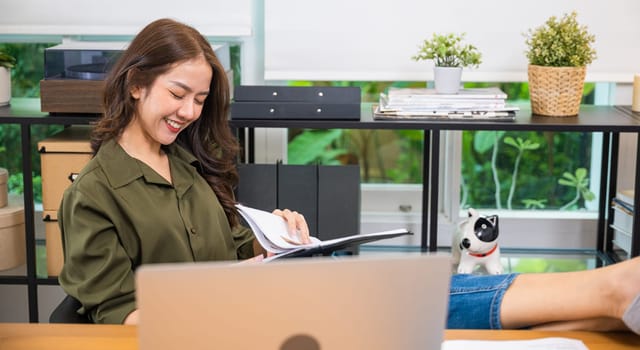 Working on financial. Businesswoman sitting relaxing reclining with feet up on computer desk reading report document in paperwork clipboard, smiling woman holding papers document reading with laptop