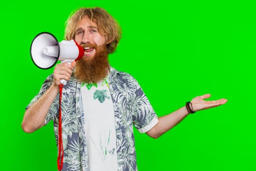 Happy caucasian man talking with megaphone, proclaiming news, loudly announcing advertisement discounts sale, using loudspeaker to shout speech. Redhead guy isolated on green chroma key background