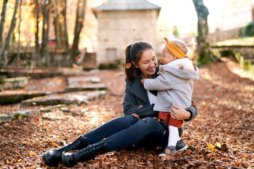Laughing mom and little girl hugging while sitting in the park under falling leaves. High quality photo