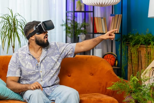 Indian man using virtual reality futuristic technology VR app headset helmet to play simulation 3D 360 online video game, watching film movie at modern home apartment. Guy in goggles sitting on sofa