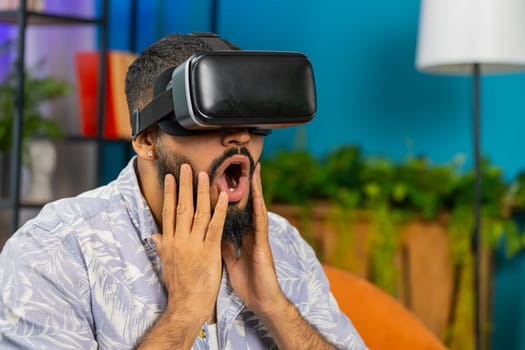 Indian man using virtual reality futuristic technology VR app headset helmet to play simulation 3D 360 online video game, watching film movie at modern home apartment. Guy in goggles sitting on sofa