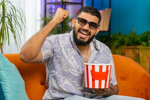 Excited Indian man sitting on sofa eating popcorn and watching interesting tv serial, sport game, film, online social media movie content at home apartment. Guy enjoying domestic entertainment indoors