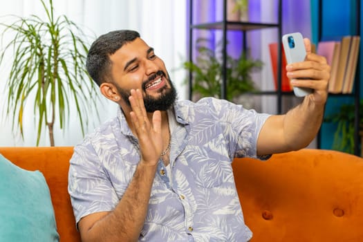 Happy Indian man blogger taking selfie on smartphone, communicating video call online with subscribers, recording stories for social media. Guy at modern home apartment living room sitting on sofa