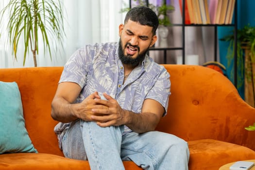 Indian man suffer from painful discomfort severe knee joint ache, leg muscles, could not walk. Sick Arabian guy massaging knees illness cramps rheumatism at home room on sofa. Health care problem