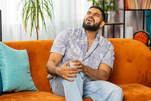 Indian man suffer from painful discomfort severe knee joint ache, leg muscles, could not walk. Sick Arabian guy massaging knees illness cramps rheumatism at home room on sofa. Health care problem