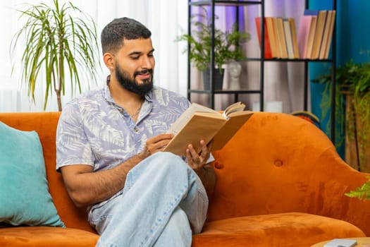 Indian man reading interesting book, turning pages smiling enjoying literature, taking a rest on comfortable couch. Portrait of peaceful cheerful guy relaxing at home apartment living room on sofa