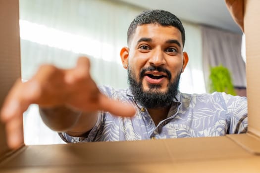 View from inside the box. Happy Indian man unpacking delivery parcel at home. Smiling satisfied guy shopper, online shop customer opening cardboard box receiving purchase gift by fast postal shipping