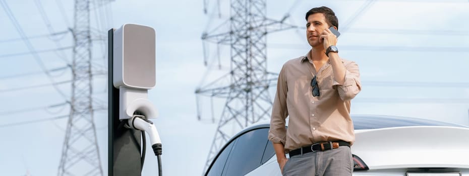 Man talking on the phone while recharge EV car battery at charging station connected to power grid tower electrical as electrical industry for eco friendly car utilization. Panorama Expedient