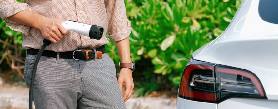 Young man recharge electric car's battery from charging station in outdoor green city park. Rechargeable EV car for sustainable environmental friendly urban travel. Panorama Expedient