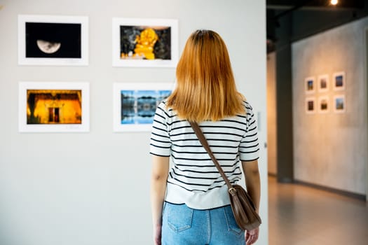 Asian woman standing she looking art gallery in front of colorful framed paintings pictures on white wall, young female watch at photo frame to leaning against at show exhibition gallery, Back view