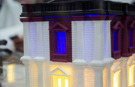 Prototype printed on 3D printer brick building with columns and glowing windows. Model of building with white and brown color, windows with light shining, created by 3D printer from molten plastic