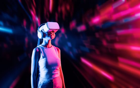 Smart female standing surrounded by neon light wearing VR headset connecting metaverse, future cyberspace community technology. Elegant woman looking faraway and smiling satisfactorily. Hallucination.