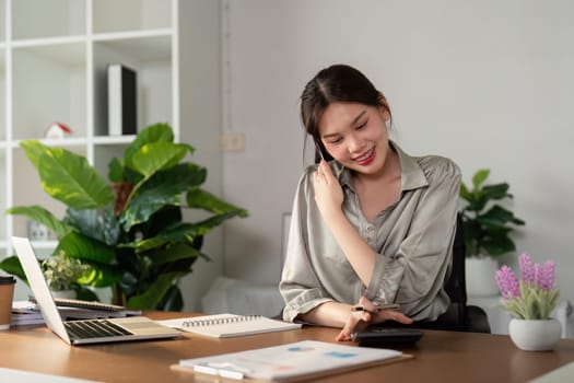 Happy smiling asian business woman working on laptop at office, using smart phone. Businesswoman sitting at office.