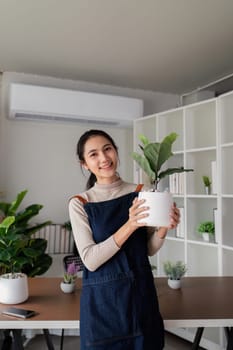 Young Asian woman smiling friendly holding flower pot with green plant house and looking in living room. Concept of home garden. Taking care of home plants.