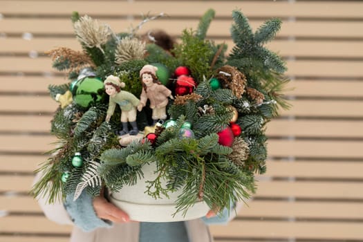 A person is shown holding a potted plant adorned with festive Christmas decorations. The individuals hand grips the pot firmly, with colorful baubles, glittering lights, and a small bow enhancing the green foliage. The background is nondescript to draw attention to the central subject.