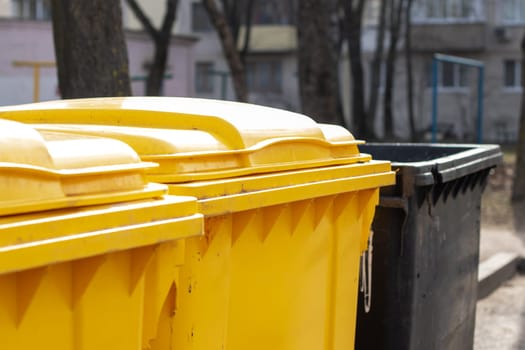 A yellow waste container made of composite material sits next to a black plastic cylinder waste containment near a wooden fence