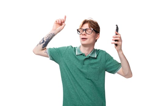 young red-haired man in a green t-shirt dancing holding a smartphone in his hand on a white background with copy space.