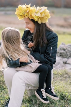 Smiling mother in wreath of yellow leaves tickles little girl sitting on her lap on lawn. High quality photo