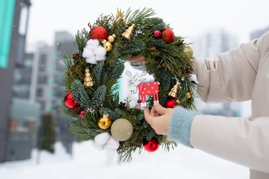 A person standing in the snow, holding a Christmas wreath with red ribbons and green foliage. The snow-covered ground contrasts with the bright colors of the wreath.