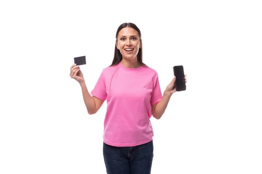 young slim brunette woman in pink basic t-shirt holding credit card mockup and smartphone on white background with copy space.