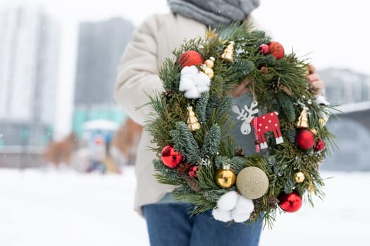 A person stands in the snow, holding a Christmas wreath. The individual is bundled up in winter clothing as they display the festive decoration.