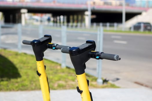 A black and yellow scooter with an electric blue bicycle handlebar is parked on the side of the asphalt road in the city, blending in with the metal car passing by. A vehicle for recreation