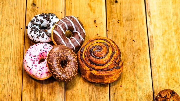 Colorful donuts, puff pastry and biscuits on wooden table. Sweet icing sugar food with glazed sprinkles, doughnut with chocolate frosting. Top view with copy space