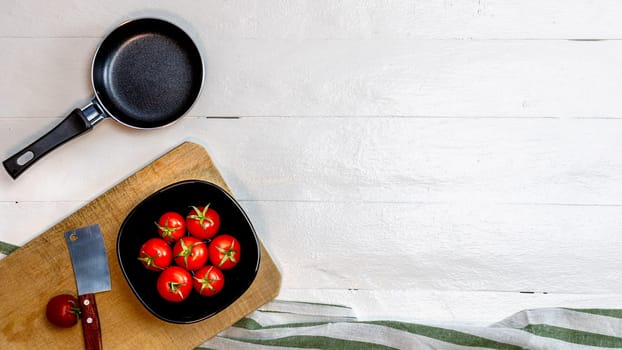 Top view of knife, small pan and fresh ripe cherry tomatoes in small black bowl on a rustic white wooden table. Ingredients and food concept