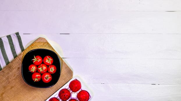 Top view of fresh ripe cherry tomatoes and red Easter eggs on a rustic white wooden table. Ingredients and food concept