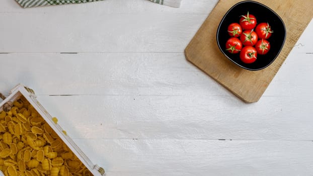 Top view of pasta in a wooden crate and fresh ripe cherry tomatoes in a black bowl on a rustic white wooden table. Ingredients and food concept