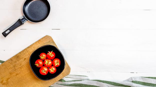 Top view of small pan and fresh ripe cherry tomatoes in small black bowl on a rustic white wooden table. Ingredients and food concept