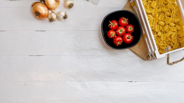 Top view of onions, garlic, pasta in a wooden crate and fresh ripe cherry tomatoes on a rustic white wooden table. Ingredients and food concept