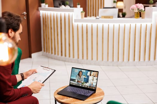 Corporate professional on videocall, using waiting time to talk to clients on remote videoconference in lounge area. Businessman attending online telework meeting in hotel lobby, business trip.