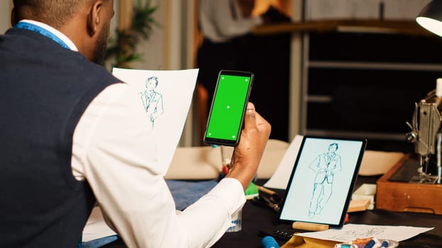 Tailor using greenscreen in atelier with team of designers, looking at isolated display on smartphone. Male couturier working with blank chroma key template on screen, tailoring concept.