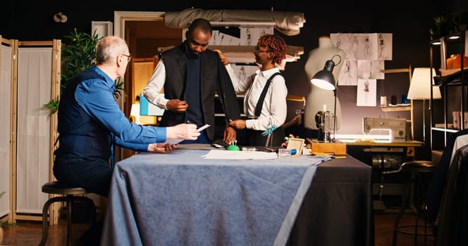 Atelier client trying on jacket to create custom made suit from experienced couturier, making sure about measurements. Senior tailor and woman manufacturing tailored blazer in workshop.