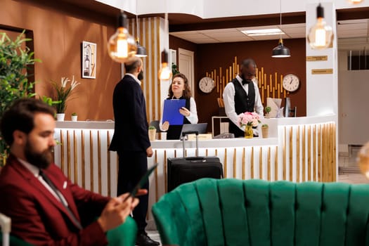 Man filling in reservation documents, approaching front desk reception with luggage in lobby. Businessman receiving good recommendations from hotel staff, travelling for work purposes.