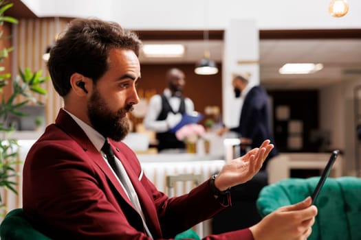 Entrepreneur attends videocall in lobby at luxury hotel, saving time before room registration. Young professional in suit using tablet to meet online with coworkers on videoconference, telework.